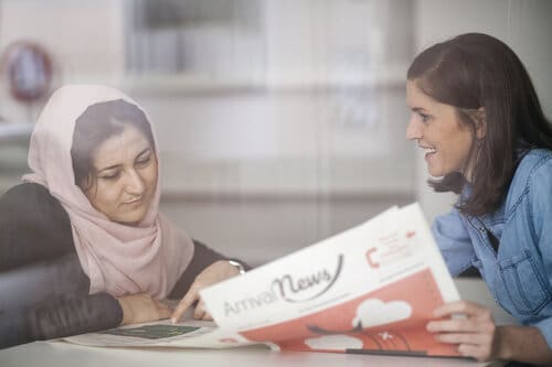 Eine Mentor*in zeigt einer Klientin eine Zeitung mit dem Titel ArrivalNews