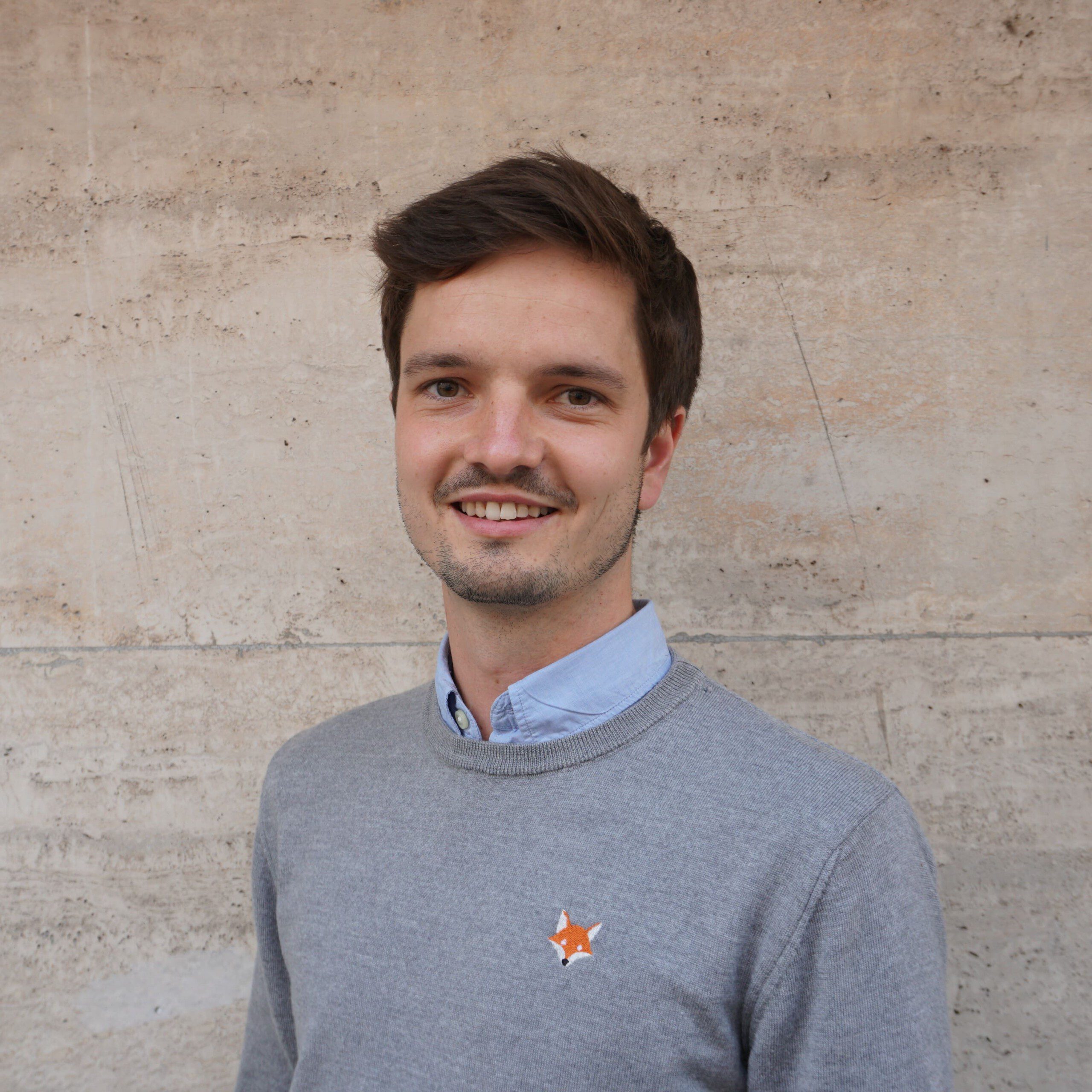 Tobias hat kurze braune Haare und einen kleinen Bart. Er trägt ein blaues Hemd und einen grauen Pullover. Er steht vor einer beigen Wand.