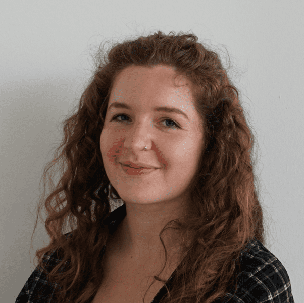 Lisi hat langes, gelocktes kastanienbraunes Haar. Sie trägt ein kariertes Hemd und steht vor einer weißen Wand.