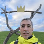 Markus hat kurze graue Haare. Er trägt einen gelben Regenmantel und einen Rucksack. Er steht vor einer Reihe von Ästen, an denen eine gelbe Krone befestigt ist. Sein Gesicht steht fast unterhalb der Krone, was den Eindruck erweckt, dass er die Krone trägt.