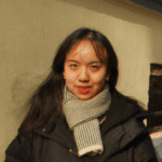 Xinran Li mit dunklen langen Haaren trägt einen grauweiß gestreiften Schal und eine schwarze Winterjacke. Sie steht vor einer gelben Mauer und lächelt leicht in die Kamera.