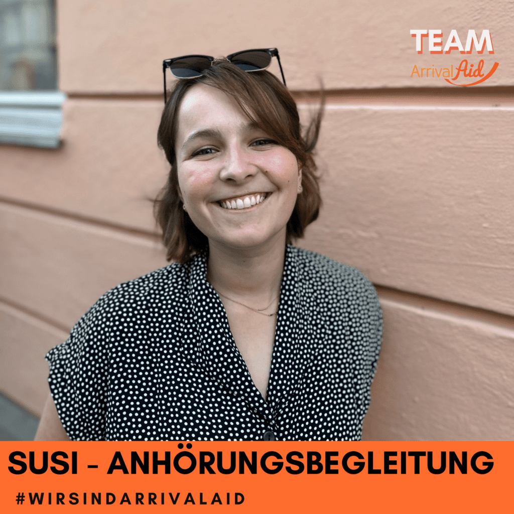 Susanne Fickenscher steht vor einer hellbraunen Wand. Sie trägt, eine schwarzweiß gepunktete Bluse, kinnlange braune haare und eine Sonnenbrille auf dem Kopf. Susi lächelt strahlend in die Kamera.
