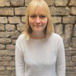 Vanessa a les cheveux blonds raides et une frange. Elle porte un pull blanc et se tient devant un mur.