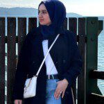 Sowaiba porte un hijab bleu, un blazer bleu et un T-shirt blanc. Elle porte un sac à main blanc sur son épaule. Elle se tient devant un lac.