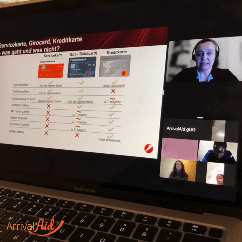 A laptop screen shows an online presentation with a table that explains the differences between service cards, debit cards and credit cards. At the top of the screen you can see a speaker speaking via video calling software, with a headset and a friendly facial expression. In the lower right area you can see other participants in the online meeting who are listening attentively. The &#39; ArrivalAid &#39; logo can be seen at the bottom left.