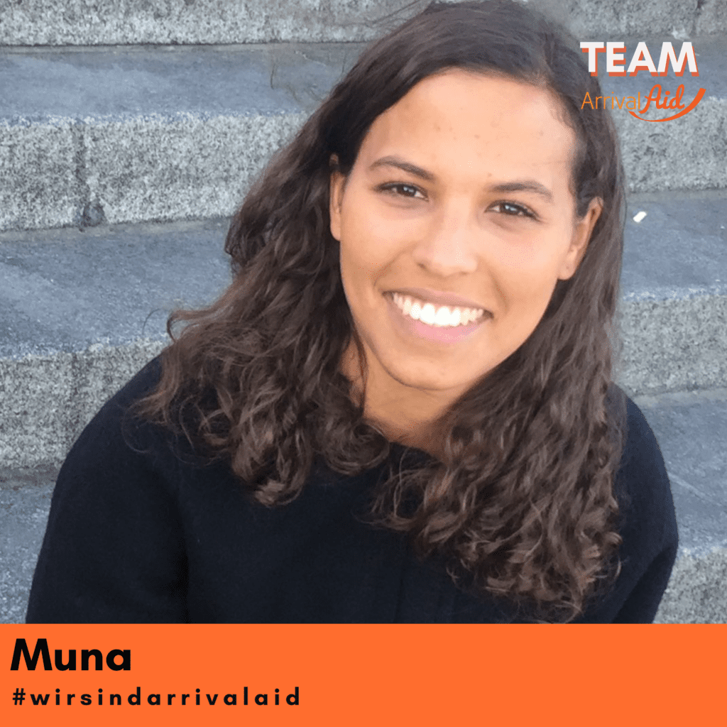 Muna Osman sits on dark gray stone steps. She has long brown curly hair. She is wearing a black sweater and smiling at the camera. In the upper right corner of the image there is a logo with the text "TEAM ArrivalAid ". At the bottom left of the picture is the name "Muna" and the hashtag "#wirsind arrivalaid " on an orange background. The picture appears friendly and positive.