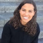 Muna Osman sits on dark gray stone steps. She has long brown curly hair. She is wearing a black sweater and smiling at the camera.