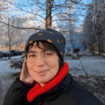 Margherita (Anfang 20) hat dunkelbraune Haare trägt eine dunkelgraue Mütze mit gelben Punkten, eine silberne runde Brille, einen roten Schal und eine schwarze Jacke. Im Hintergrund sind man Bäume und Schnee. Sie lächelt.