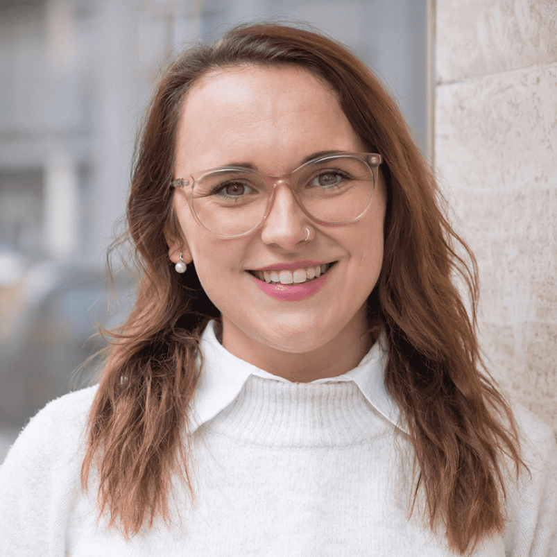 Alena trägt eine Brille, hat schulterlanges mittelbraunes Haar. Sie trägt ein weißes Hemd und einen Pullover darüber.