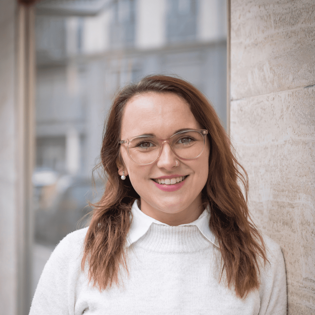 Alena trägt eine Brille, hat schulterlanges mittelbraunes Haar. Sie trägt ein weißes Hemd und einen Pullover darüber.