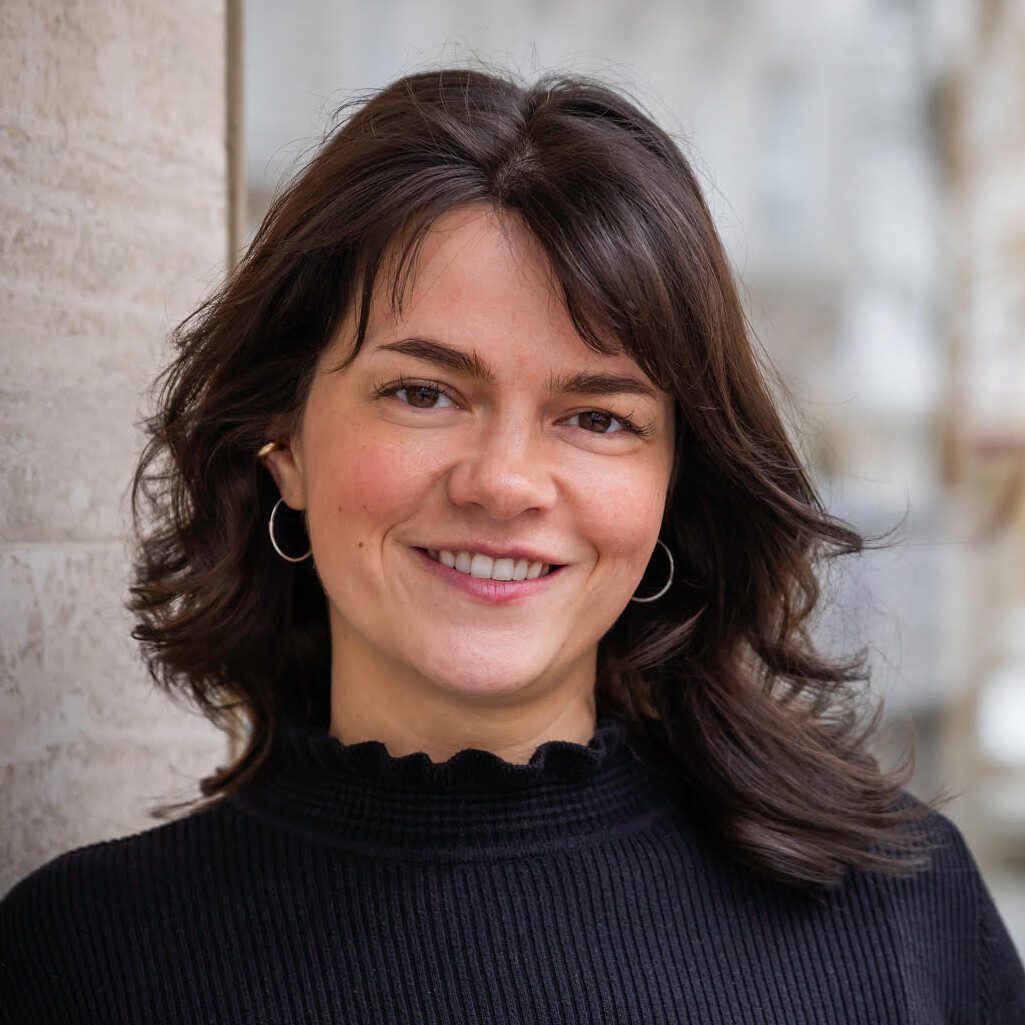 Katharina Stoib Frau mit braunen welligen, Schulterlangen Haaren. Sie trägt einen schwarzen Pullover mit welligen Kragen und lächelt.