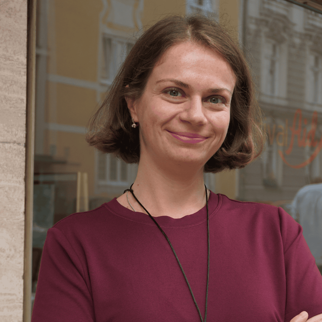 Anna Shapiro hat heller Haut und kurzen, braunen Haare. Sie trägt ein bordeauxrotes Oberteil und eine lange schwarze Kette.